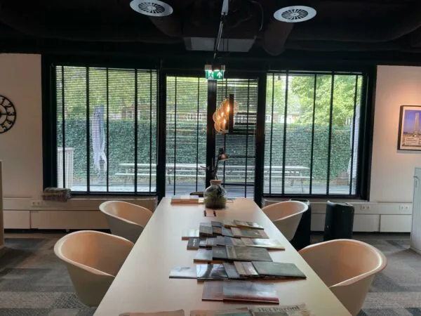 Moderne vergaderruimte aan de De Cuserstraat met een lange tafel, designstoelen en tijdschriften, met uitzicht op een groene tuin door grote ramen met jaloezieën.